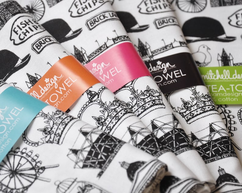 Może przedstawiać: Zestaw pięciu ręcznik&oacute;w kuchennych z czarno-białymi ilustracjami londyńskich zabytk&oacute;w, w tym London Eye, Big Ben i Tower Bridge. Ręczniki są białe z czarnym tekstem, kt&oacute;ry brzmi "Fish Chips Brick Lane" i "Mitchell Design Tea Towels Handmade Cotton".