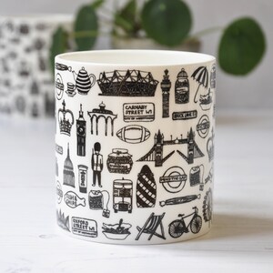 May include: White ceramic mug with a black and white design featuring iconic London landmarks, including Big Ben, the London Eye, and Tower Bridge. The mug also features text such as "Carnaby Street W1", "Brick Lane E1", and "Oxford Street W1".
