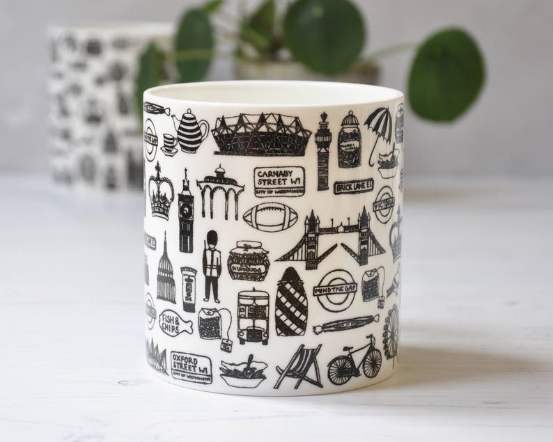 May include: White ceramic mug with a black and white design featuring iconic London landmarks, including Big Ben, the London Eye, and Tower Bridge. The mug also features text such as "Carnaby Street W1", "Brick Lane E1", and "Oxford Street W1".