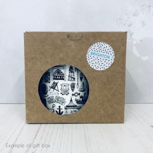 May include: A brown cardboard gift box with a white circle sticker that says "BRIGHTON" in black text. The box has a cut-out window that shows a white mug with a black and white illustration of Brighton landmarks.