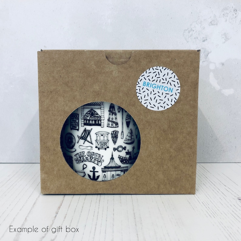 May include: A brown cardboard gift box with a white circle sticker that says "BRIGHTON" in black text. The box has a cut-out window that shows a white mug with a black and white illustration of Brighton landmarks.