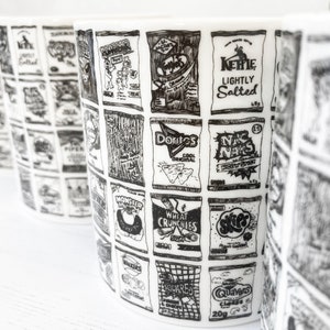 May include: A white ceramic mug with a black and white illustration of various snack food bags. The bags feature names like "Doritos", "Monster", "Wheat Crunchies", "Kehle Lightly Salted", "NK Naks", "Walkers Squares", and "Quavers Cheese".