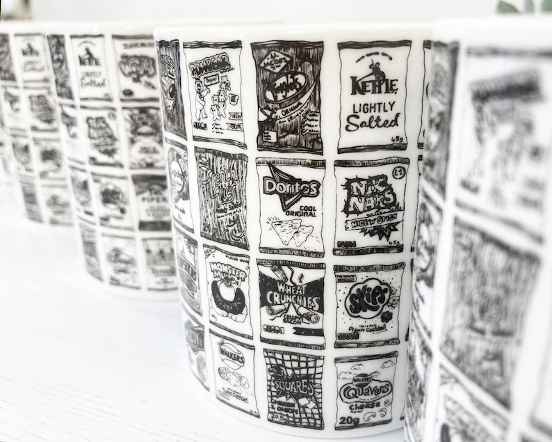 May include: A white ceramic mug with a black and white illustration of various snack food bags. The bags feature names like "Doritos", "Monster", "Wheat Crunchies", "Kehle Lightly Salted", "NK Naks", "Walkers Squares", and "Quavers Cheese".