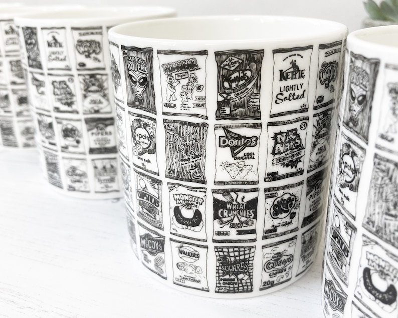 May include: A white ceramic mug with a black and white design of various snack food bags. The bags include brands like Monster Munch, McCoy's, Walkers, and Doritos.