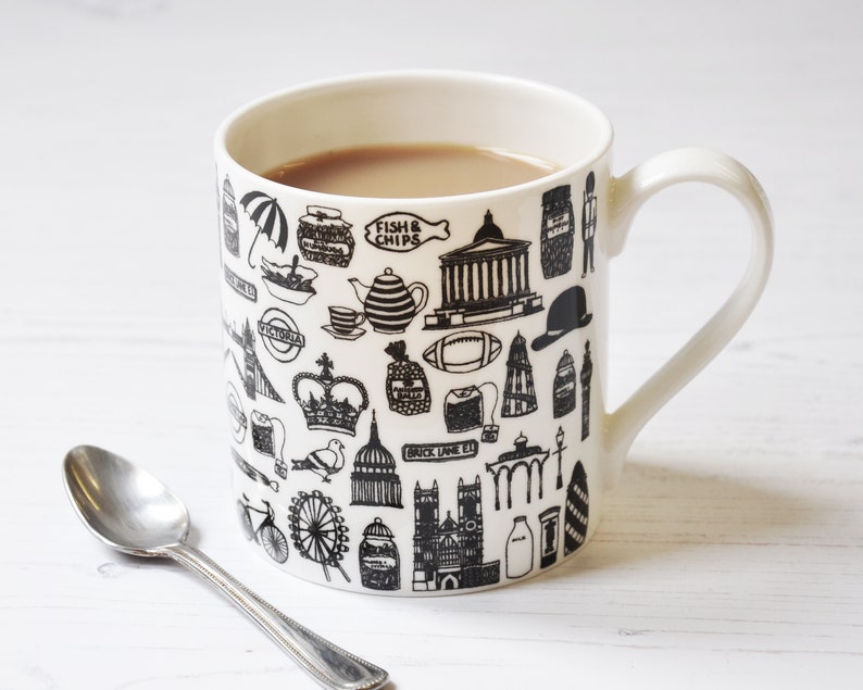 May include: A white ceramic mug with a black and white design featuring iconic London landmarks, including Big Ben, the London Eye, and a double-decker bus. The mug is filled with a brown liquid, likely tea or coffee.