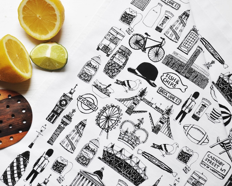 Może przedstawiać: Biały ręcznik do herbaty z czarno-białą ilustracją londyńskich zabytk&oacute;w i symboli. Projekt zawiera London Eye, Big Ben, autobus piętrowy, czerwoną budkę telefoniczną, koronę oraz napisy "VICTORIA", "FISH & CHIPS", "BRICK LANE E1" i "CARNABY STREET W1".