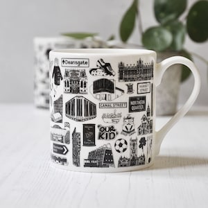 Puede incluir: Taza de cerámica blanca con una ilustración en blanco y negro de puntos de referencia y texto de Manchester, Inglaterra. El texto incluye "Canal Street", "Northern Quarter", "Our Kid" y "Ship Canal".