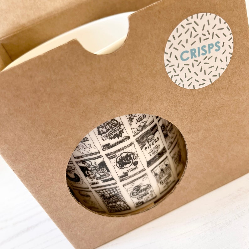 May include: A brown cardboard box with a white ceramic mug inside. The mug has a black and white print of various British crisp packets. The box has a white circle sticker with the word "CRISPS" in blue text.