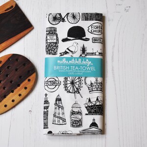 Może przedstawiać: Czarno-biały ręcznik do herbaty z motywem Londynu. Wz&oacute;r przedstawia ikoniczne zabytki, takie jak London Eye, Big Ben i piętrowy autobus. Tekst na ręczniku brzmi "FISH & CHIPS", "BRICK LANE", "VICTORIA" i "BRITISH TEA-TOWEL".
