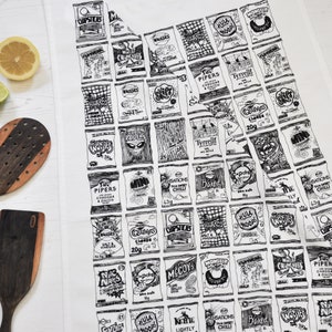 Op de afbeelding: Een witte theedoek met zwart-wit illustraties van verschillende populaire Britse chipsmerken. De merken omvatten Walkers, McCoy's, Sensations, Hula Hoops en meer.