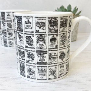 May include: White ceramic mug with a black and white illustration of various chip bag designs. The designs include the text "Kettle Lightly Salted", "Walkers Salt & Vinegar", "Skips", "Pipers Cheddar & Onion", "Sensations Thai Sweet Chilli", "Salt Shake", "Quavers Cheese 20g", "Tyrrells", "Frozies", and "Walkers Crisps".