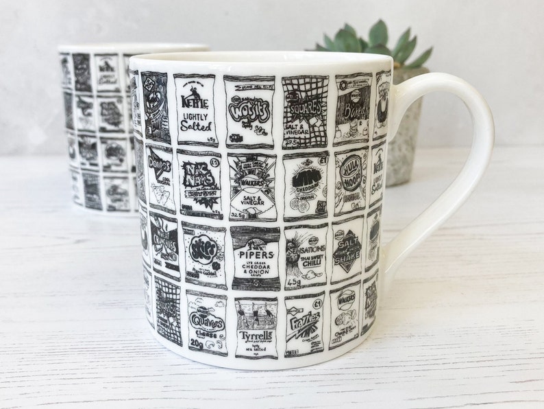May include: White ceramic mug with a black and white illustration of various chip bag designs. The designs include the text "Kettle Lightly Salted", "Walkers Salt & Vinegar", "Skips", "Pipers Cheddar & Onion", "Sensations Thai Sweet Chilli", "Salt Shake", "Quavers Cheese 20g", "Tyrrells", "Frozies", and "Walkers Crisps".