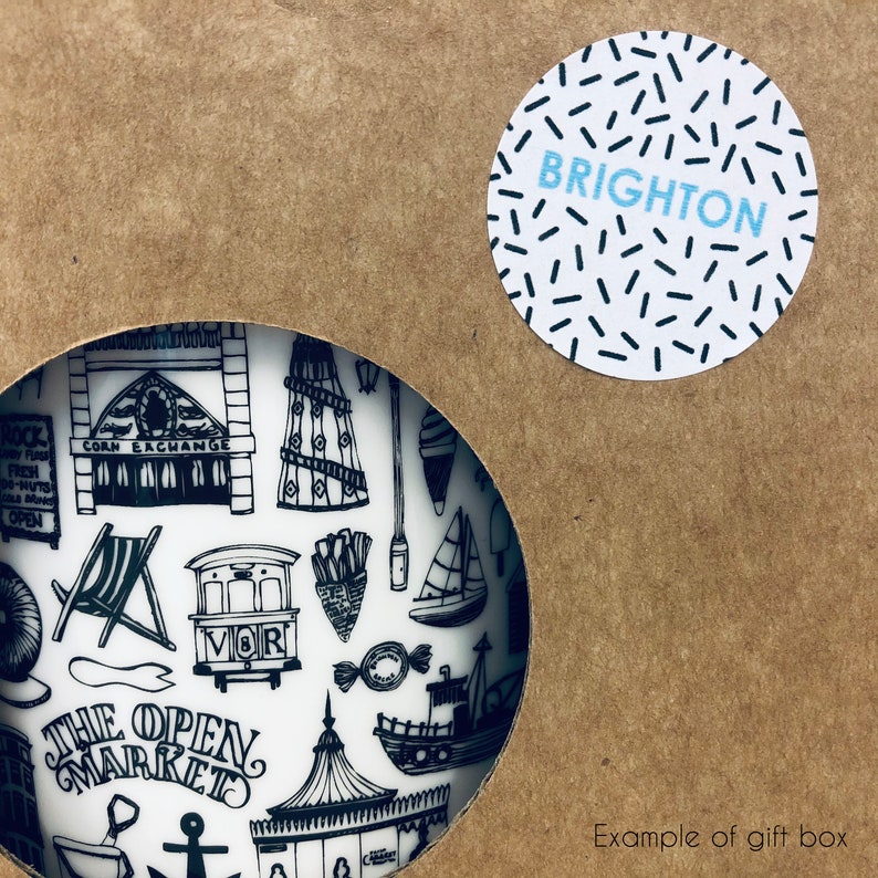 May include: A white ceramic mug featuring a black and white illustration of various Brighton landmarks, including the Corn Exchange, the Brighton Pier, and the Brighton Palace Pier. The mug is packaged in a brown cardboard box with a white circle sticker that says "BRIGHTON" in black text.