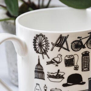 May include: White ceramic mug with a black and white design featuring iconic London landmarks, including the London Eye, Big Ben, a red double-decker bus, a black cab, and a red telephone box.