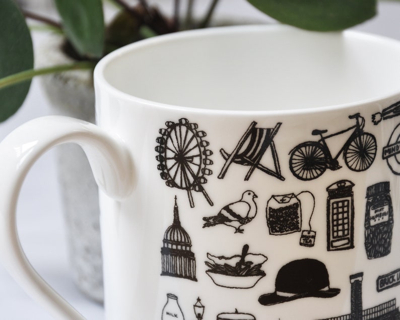 May include: White ceramic mug with a black and white design featuring iconic London landmarks, including the London Eye, Big Ben, a red double-decker bus, a black cab, and a red telephone box.