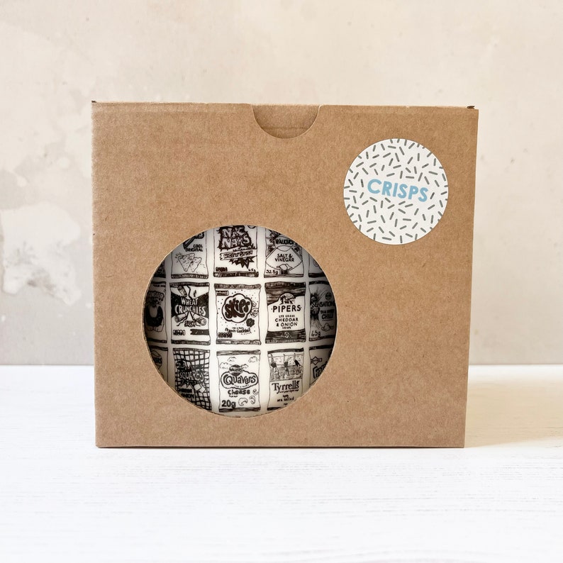 May include: A brown cardboard box with a white circle sticker that says "CRISPS" in blue text. The box has a cut-out window that shows a white mug with a black and white pattern of vintage crisp packets.