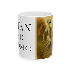 Puede incluir: Taza de cerámica blanca con el texto "EN D OMO" en negro. La taza presenta una ilustración de estilo vintage de dos extraterrestres verdes en un campo con una casa al fondo. Los extraterrestres llevan túnicas de color claro.