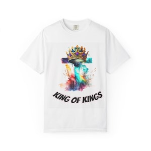 Puede incluir: Camiseta blanca con un diseño de acuarela de una cruz, una corona y acentos coloridos. Las palabras "KING OF KINGS" están impresas en una fuente negra curva en la parte inferior del diseño.