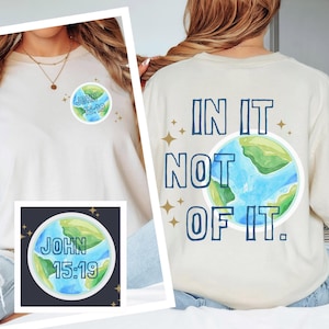 May include: A cream-coloured t-shirt featuring a watercolour design of the Earth and the text "JOHN 15:19" on the front. The back of the shirt displays the words "IN IT NOT OF IT" above a similar Earth graphic.