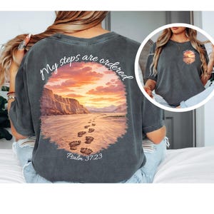 Op de afbeelding: Donkergrijs t-shirt met een landschapsontwerp. Het heeft een zonsondergang, voetstappen en de tekst "My steps are ordered" en "Psalm 37:23". Het ontwerp omvat een landschap met een pad en een kleurrijke lucht.
