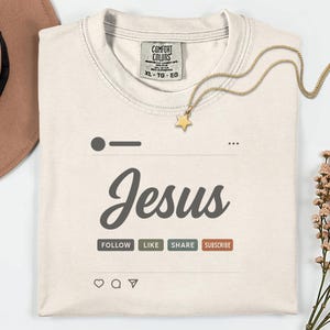 Op de afbeelding: Crèmekleurig T-shirt met het woord "Jesus" in een gestileerd lettertype, ontworpen om op een social media post te lijken. Het shirt bevat de knoppen "Follow", "Like", "Share" en "Subscribe". Een gouden sterrenketting ligt op het shirt.
