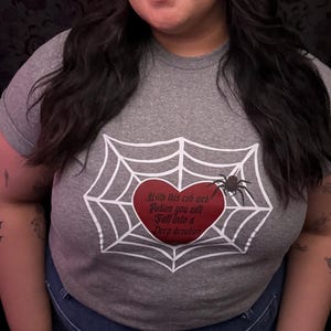 May include: A heather gray t-shirt featuring a white spider web design with a red heart at the center. The heart contains the text "With this cob web before you will fall into a deep slumber" and a black spider.
