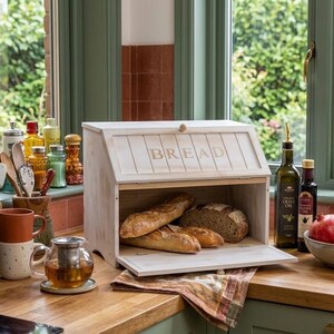 May include: A rectangular wooden bread box with a hinged front door featuring the word BREAD in capital letters. The box has a light, distressed finish and contains two long loaves of bread and one round loaf of bread inside the storage compartment.