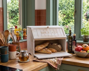 May include: A rectangular wooden bread box with a hinged front door featuring the word BREAD in capital letters. The box has a light, distressed finish and contains two long loaves of bread and one round loaf of bread inside the storage compartment.