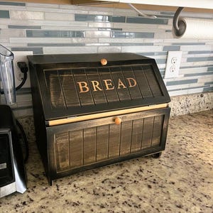 May include: A black wooden bread box with the word "BREAD" in copper-colored letters on the front. The box has a hinged lid and a wooden knob. The bread box is sitting on a granite countertop.