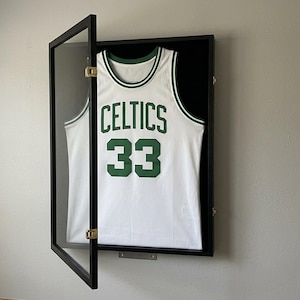 May include: A white Boston Celtics basketball jersey with the number 33 in green, displayed inside a black rectangular wall-mounted display case with a hinged glass door and a black interior backing to showcase the sports memorabilia.
