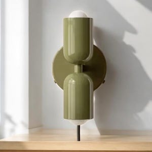 May include: A green wall-mounted light fixture with two cylindrical shades. The shades have a white interior and are connected by a black metal rod.