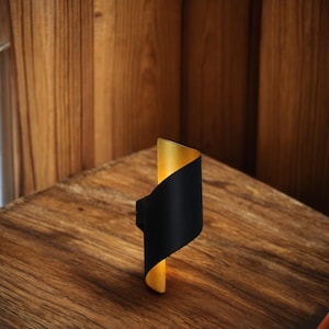 Può includere: Un'applique da parete moderna nera e dorata con un design curvo. L'applique è accesa e proietta una luce calda.