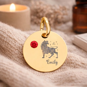 以下が含まれることがあります： 赤い宝石と獅子座のサインの彫刻が施された、金色の丸いペンダント。ペンダントの上部にはループがあり、下部には「Emily」という名前が刻まれています。ペンダントはベージュ色の表面にあります。