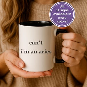 May include: White ceramic mug with a black interior and handle. The mug features the text "can't i'm an aries" in black. A purple circle with white text that says "All 12 signs available in more colors!" is also visible.