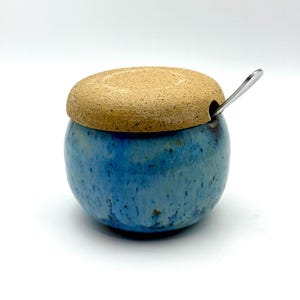 May include: A blue ceramic sugar bowl with a speckled tan lid and a small stainless steel spoon. The bowl has a round shape and a glossy finish with shades of blue. The lid sits on top of the bowl, and the spoon is inserted into a small hole.