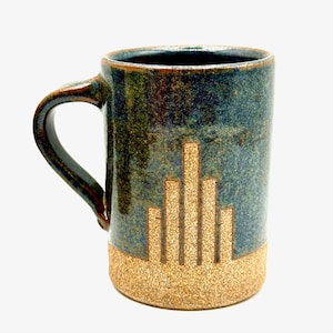 May include: A handmade ceramic mug with a dark blue and brown glaze. The mug features a textured, tan-colored base and a vertical, geometric design in a lighter tan color. The handle is brown.