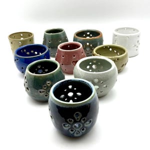May include: A collection of ten small, round ceramic vessels in various colors including blue, green, pink, white, and brown. Each vessel features a series of circular cutouts arranged in patterns around the exterior and base of the object.