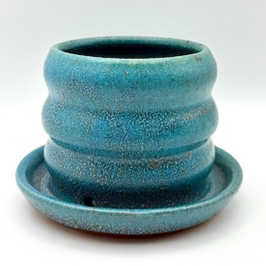 May include: A cylindrical ceramic planter with a vibrant turquoise glaze and a speckled texture. The planter features three horizontal ridges along its body and sits on a matching circular saucer with a small drainage hole visible at the base.