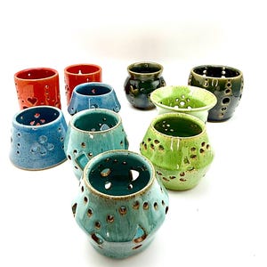 May include: A collection of nine ceramic candle holders in various colors including red, blue, teal, green, and dark olive. Each vessel features a glazed finish and numerous circular cutouts of different sizes arranged in patterns around the body.