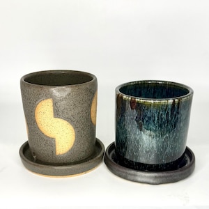 May include: Two ceramic planters with matching saucers. One is gray with yellow geometric shapes, the other is dark blue-green with a glossy finish. Both are cylindrical and suitable for small plants.