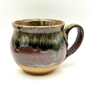 May include: Handmade ceramic mug with a rounded shape and a dark handle. The mug features a textured, earthy design with a blend of brown, green, and burgundy colors. The base has a natural, unglazed appearance.