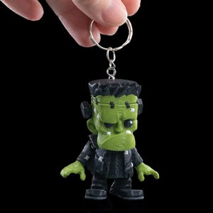 May include: A green Frankenstein's monster keychain with a silver keyring. The figure has a textured face, black hair, and a black jacket. The keychain is held against a black background.