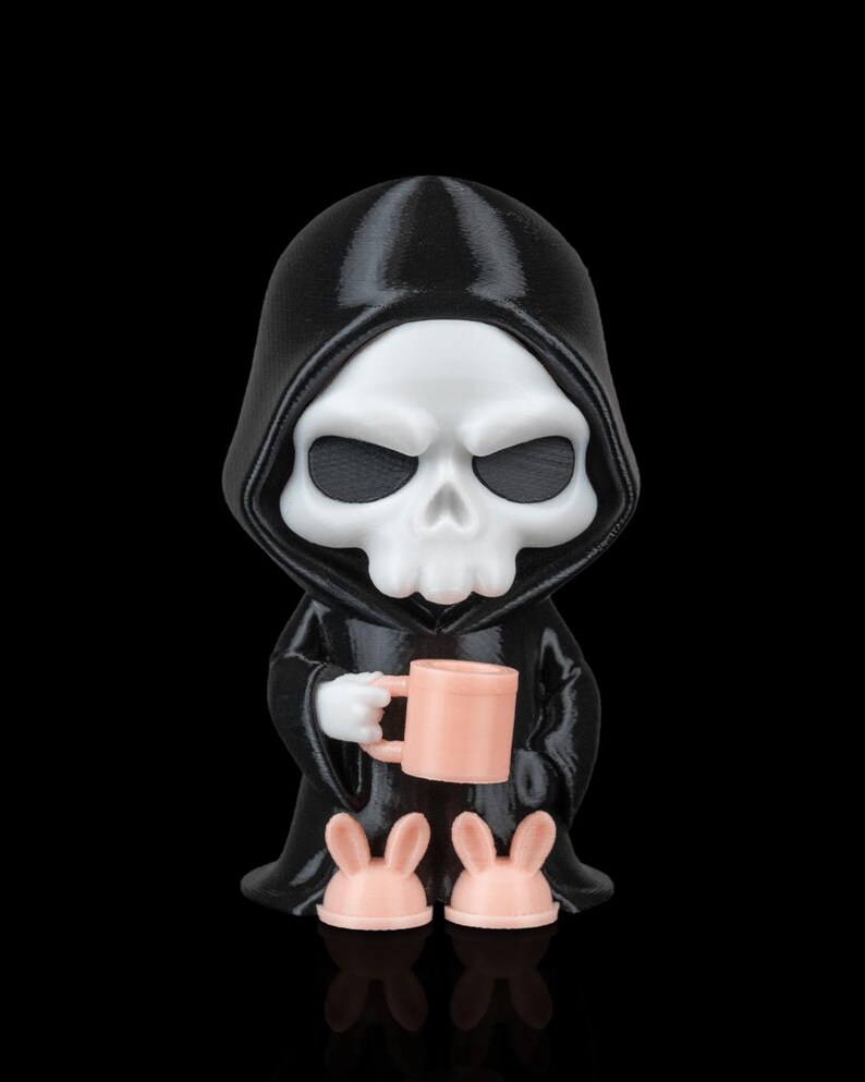 K&ouml;nnte beinhalten: 3D-gedruckte Figur des Sensenmanns in einer schwarzen Kapuzenrobe, die eine rosa Tasse h&auml;lt und rosa Hasenpantoffeln tr&auml;gt. Die Figur hat ein wei&szlig;es Totenkopfgesicht und ist vor schwarzem Hintergrund platziert.