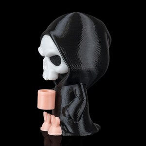 K&ouml;nnte beinhalten: Eine 3D-gedruckte Figur des Sensenmanns, in einem schwarzen Kapuzenmantel, der einen rosa Becher und zwei rosa Hasenfiguren h&auml;lt. Der Sch&auml;del ist wei&szlig;, vor schwarzem Hintergrund.