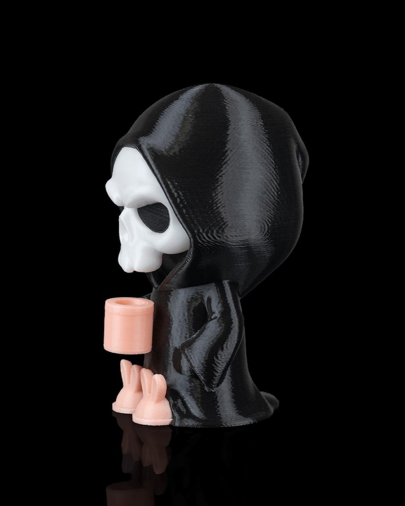K&ouml;nnte beinhalten: Eine 3D-gedruckte Figur des Sensenmanns, in einem schwarzen Kapuzenmantel, der einen rosa Becher und zwei rosa Hasenfiguren h&auml;lt. Der Sch&auml;del ist wei&szlig;, vor schwarzem Hintergrund.