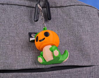 May include: A Halloween-themed keychain featuring a pumpkin-headed dinosaur. The orange pumpkin head has black eyes and a smile. The dinosaur body is green and tan. The keychain is attached to a gray backpack.