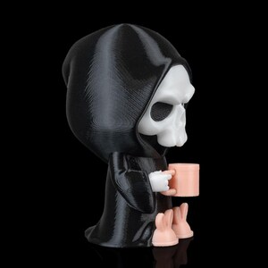 K&ouml;nnte beinhalten: Eine schwarz-wei&szlig;e Figur des Sensenmanns, der einen rosa Becher h&auml;lt. Die Figur hat eine schwarze Kapuzenrobe, ein wei&szlig;es Totenkopfgesicht und rosa Hasenf&uuml;&szlig;e. Der Becher ist ebenfalls rosa. Die Figur steht auf einer schwarzen Oberfl&auml;che.
