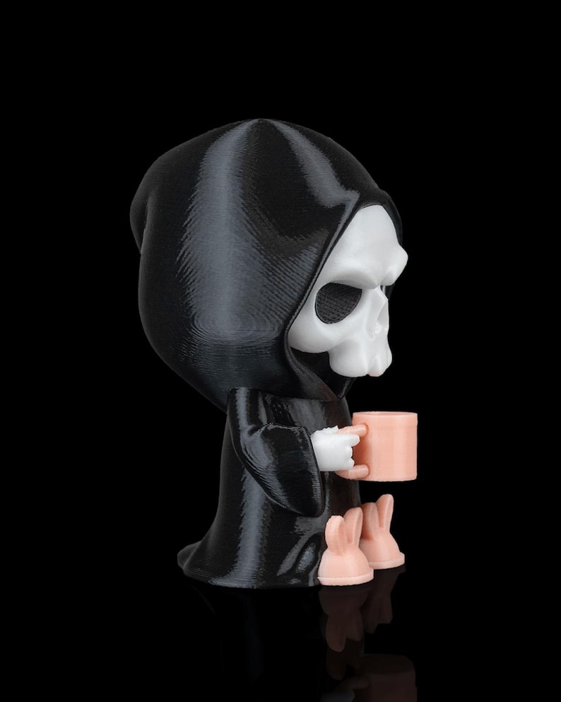 K&ouml;nnte beinhalten: Eine schwarz-wei&szlig;e Figur des Sensenmanns, der einen rosa Becher h&auml;lt. Die Figur hat eine schwarze Kapuzenrobe, ein wei&szlig;es Totenkopfgesicht und rosa Hasenf&uuml;&szlig;e. Der Becher ist ebenfalls rosa. Die Figur steht auf einer schwarzen Oberfl&auml;che.