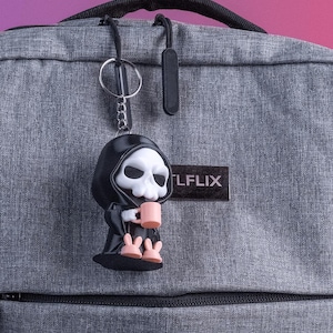 K&ouml;nnte beinhalten: Ein schwarzer Schl&uuml;sselanh&auml;nger mit einer Sensemann-Figur mit wei&szlig;em Totenkopfgesicht, die einen rosa Becher h&auml;lt. Der Schl&uuml;sselanh&auml;nger ist an einem grauen Rucksack befestigt. Der Rucksack hat einen schwarzen Rei&szlig;verschluss und das Wort "LFLIX" auf einem schwarzen Aufn&auml;her.
