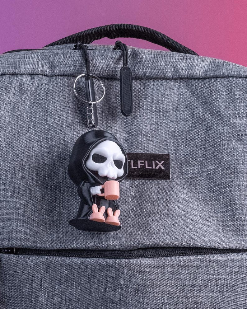 K&ouml;nnte beinhalten: Ein schwarzer Schl&uuml;sselanh&auml;nger mit einer Sensemann-Figur mit wei&szlig;em Totenkopfgesicht, die einen rosa Becher h&auml;lt. Der Schl&uuml;sselanh&auml;nger ist an einem grauen Rucksack befestigt. Der Rucksack hat einen schwarzen Rei&szlig;verschluss und das Wort "LFLIX" auf einem schwarzen Aufn&auml;her.